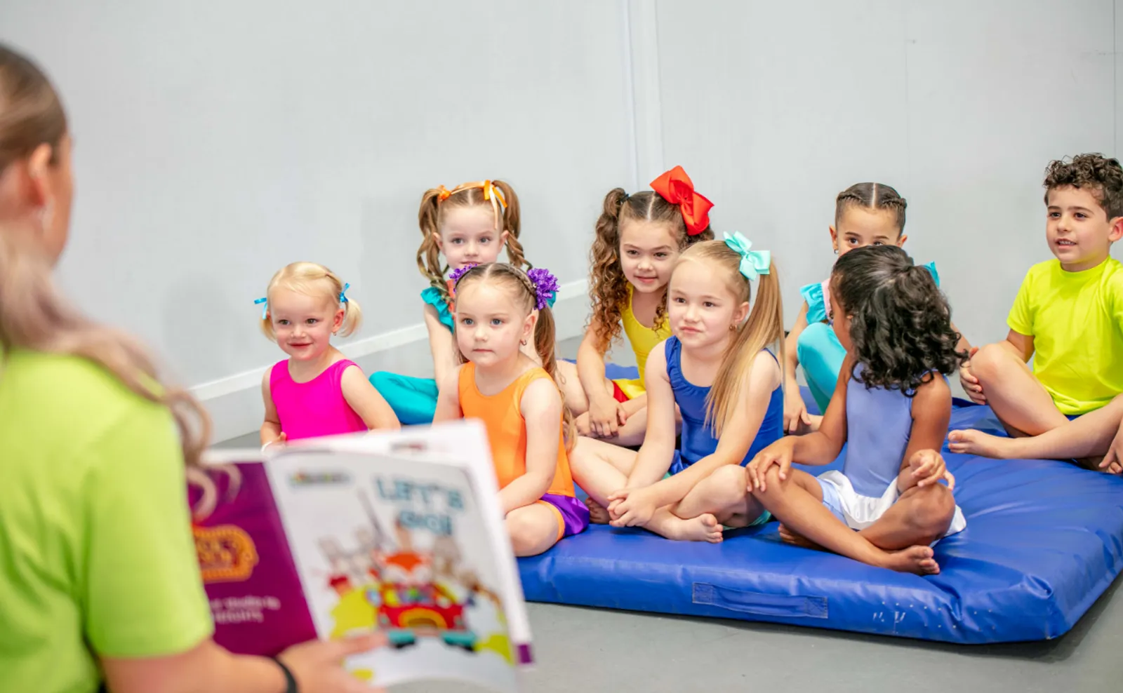 Young children in colourful leotards on a gym mat, listening to their teacher read a picture book in class