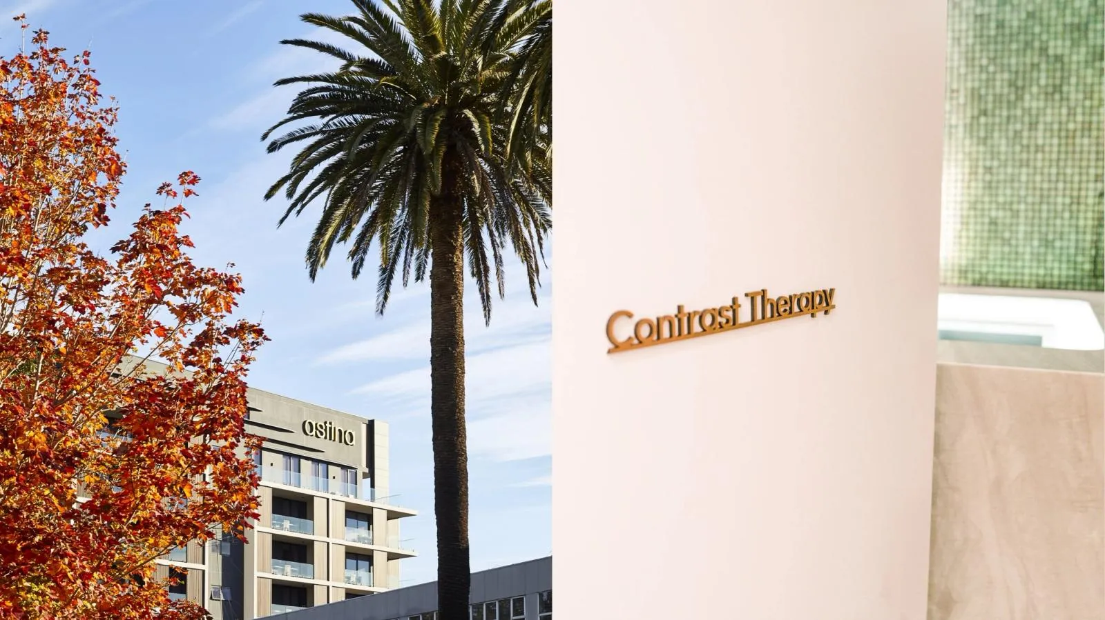 Astina Suites tower framed by autumn leaves and a palm, beside brass "Contrast Therapy" wayfinding