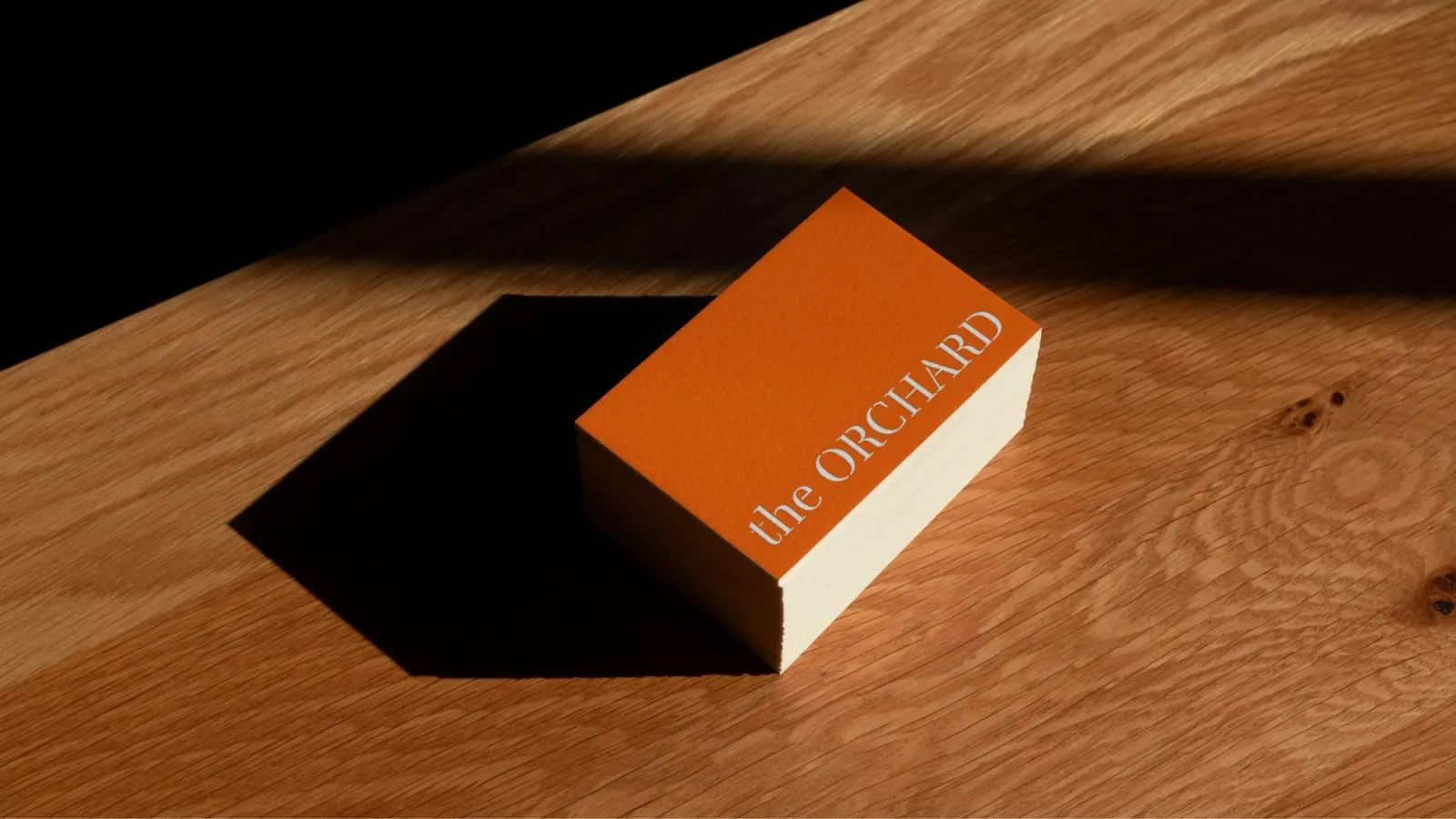 A stack of orange The Orchard business cards on a timber table in afternoon light