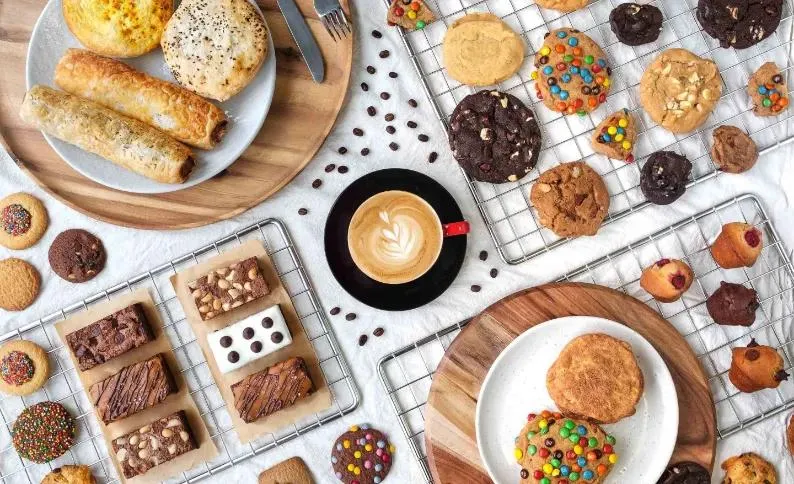 Flat lay of Mrs Fields cookies, brownies, muffins and sausage rolls around a cup of coffee