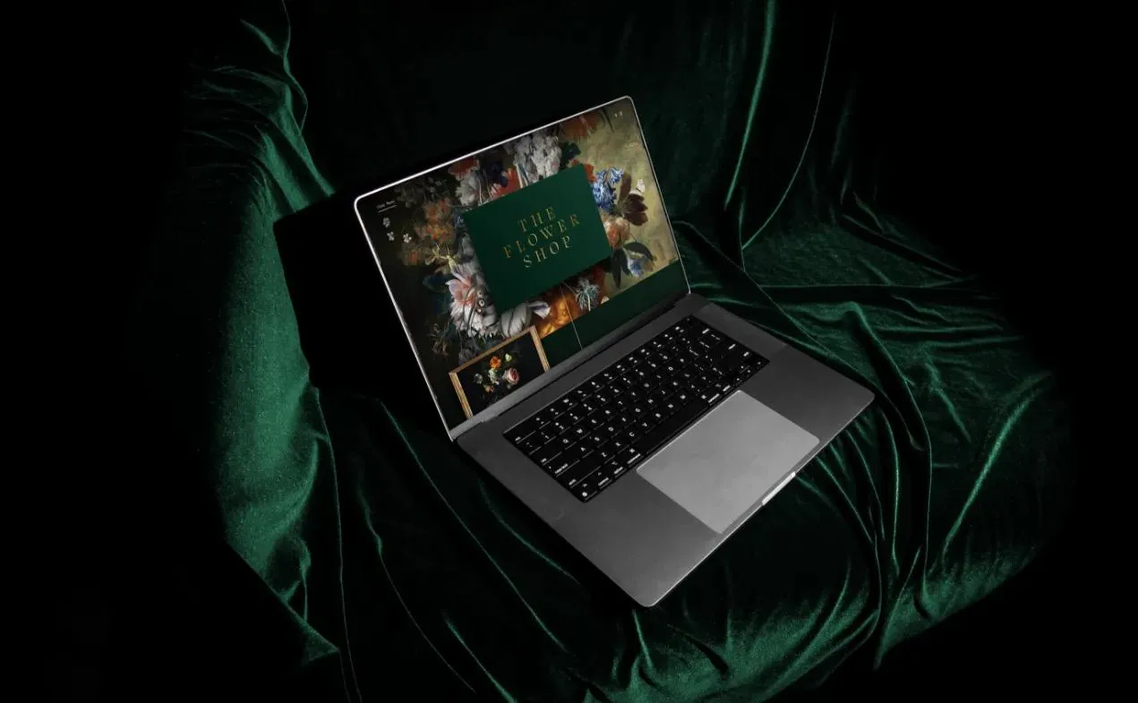 The Flower Shop website on a laptop resting on green velvet, its floral homepage framing the gold logo