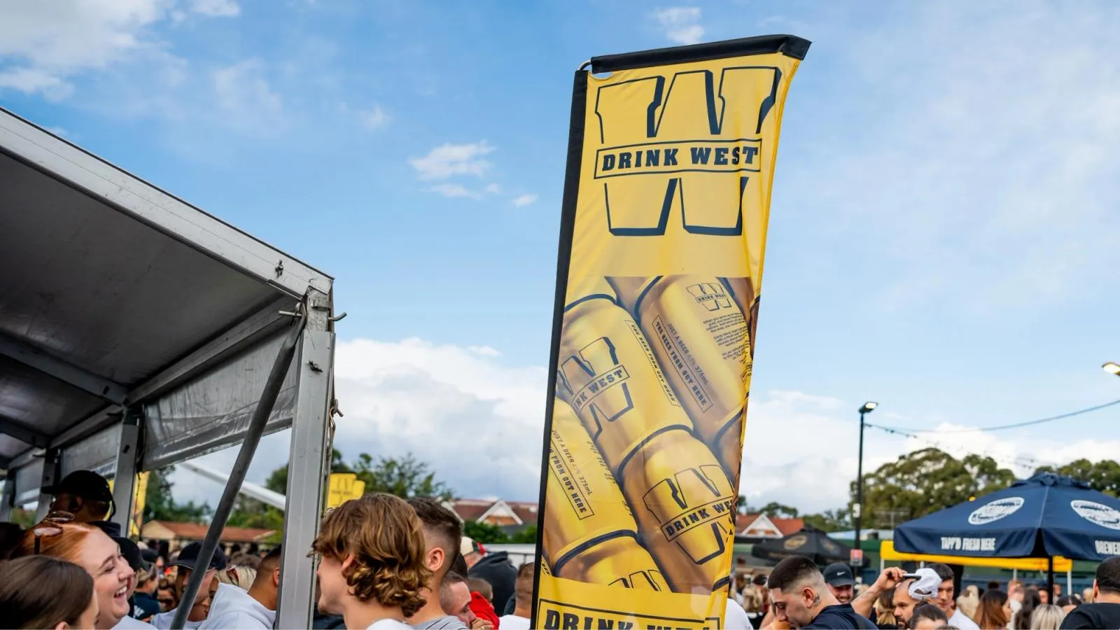 Drink West feather flag flying above a crowd at an outdoor event marquee on a sunny day