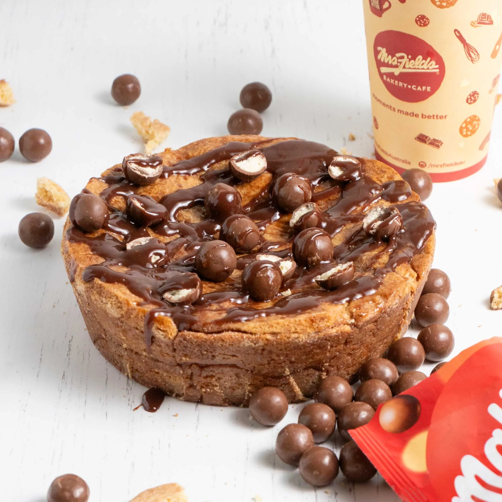 Mrs Fields cookie cake topped with Maltesers, beside a Mrs Fields cup